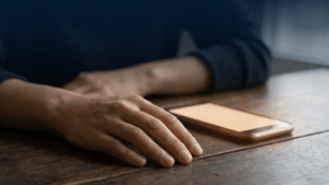 Eine entspannte Hand liegt auf einem dunklen Holztisch neben einem flach liegenden Smartphone, dessen Display warm und ohne Text leuchtet, vor einem ruhigen, dunklen Hintergrund.